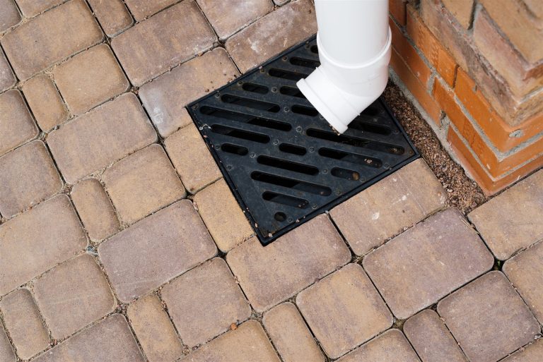 White downspout emptying into a drain grate set in brick pavers
